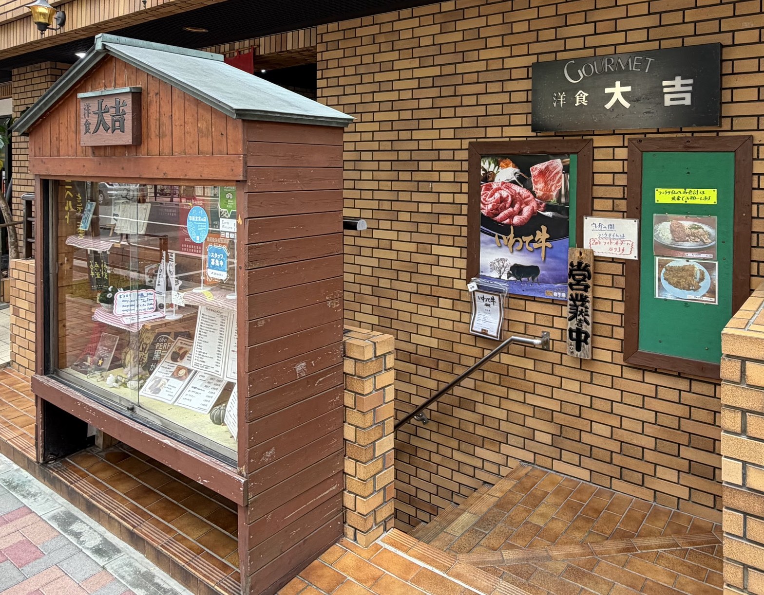 レンガ壁に「GOURMET 洋食 大吉」の黒い看板、手前に木製のメニューショーケース、「営業中」の札と料理写真が貼られた入口