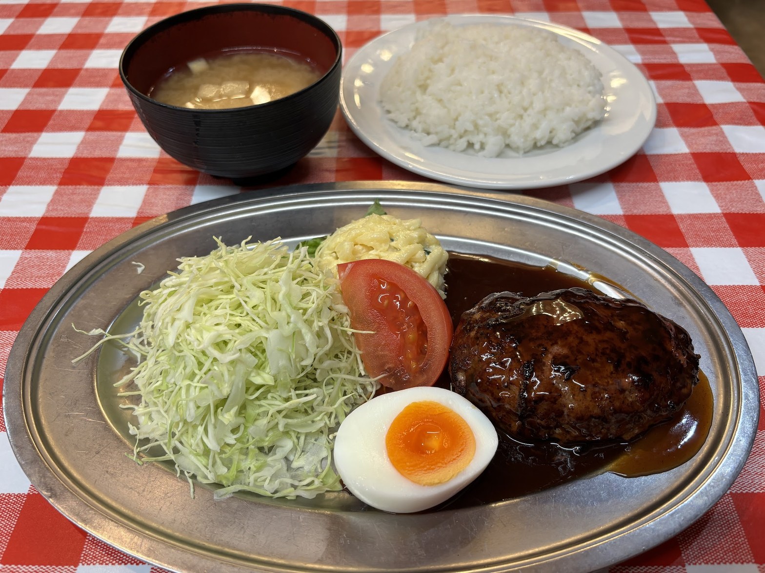 赤白チェックのテーブルクロスの上に置かれたステンレス楕円皿のハンバーグ定食。黒いソースがかかったハンバーグ、半熟ゆで卵、千切りキャベツ、トマト、奥に味噌汁と白飯