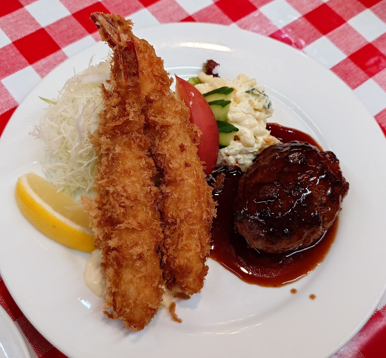 白い丸皿に盛られた大ぶりエビフライ2本とハンバーグ。エビフライにはレモンとキャベツの千切り、ハンバーグには茶色いソースがかかり赤白チェックのクロスが背景